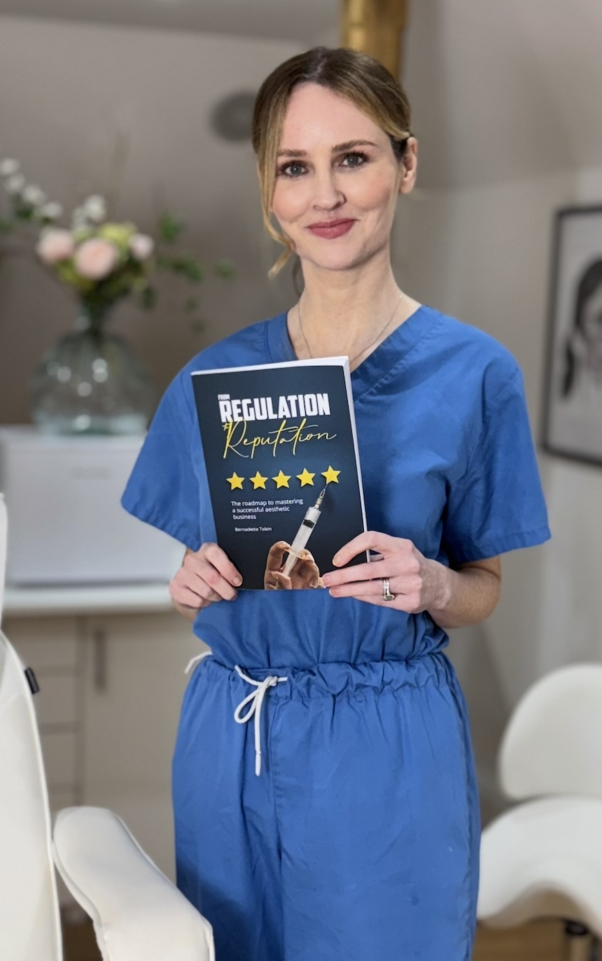 Bernadette Tobin in scrubs holding her book Regulation to Reputation in her clinic